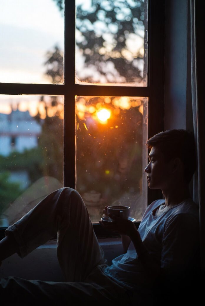 warm-toned photo of someone sitting by a window during sunset, holding a cup of tea, looking thoughtful but calm.
